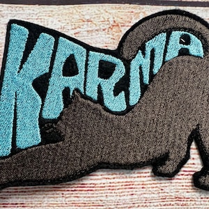 Peut inclure: Écusson brodé avec le mot "KARMA" en bleu clair, souligné de noir, et une silhouette de chat marron. Le chat est arqué, sa queue formant la lettre "A". L'écusson a une bordure noire.