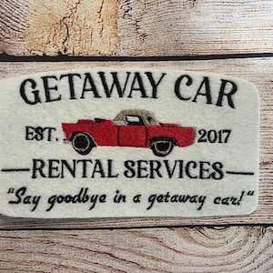 Puede incluir: Un letrero de fieltro blanco con texto bordado en negro que dice "GETAWAY CAR EST. 2017 -RENTAL SERVICES- "Say goodbye in a getaway car!" Un coche rojo antiguo est&aacute; bordado en el centro del letrero.