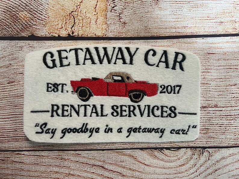 Puede incluir: Un letrero de fieltro blanco con texto bordado en negro que dice "GETAWAY CAR EST. 2017 -RENTAL SERVICES- "Say goodbye in a getaway car!" Un coche rojo antiguo est&aacute; bordado en el centro del letrero.