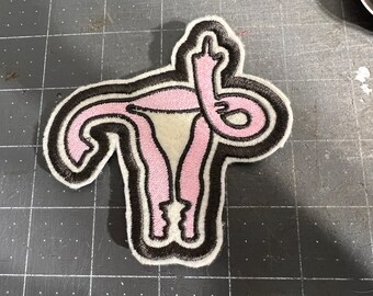 Puede incluir: Parche bordado con un diseño estilizado de útero rosa con contornos negros y blancos. El útero muestra el dedo medio. El parche está sobre una superficie gris oscuro.