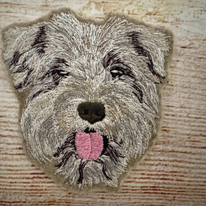 Op de afbeelding: Geborduurde patch met een honden gezicht in lichtgrijs en bruin. De hond heeft een zwarte neus, donkerbruine accenten en een roze tong. De patch is geplaatst op een lichtbruine achtergrond met houtnerf.