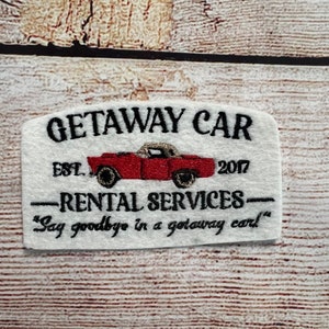 May include: White embroidered patch with the text "GETAWAY CAR EST. 2017" and "RENTAL SERVICES" with a red car illustration and the text "Say goodbye in a getaway car."
