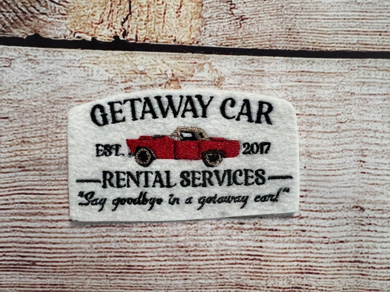 May include: White embroidered patch with the text "GETAWAY CAR EST. 2017" and "RENTAL SERVICES" with a red car illustration and the text "Say goodbye in a getaway car."