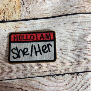 May include: A white name tag with a red top and black border. The text on the tag reads "HELLO I AM She/Her".
