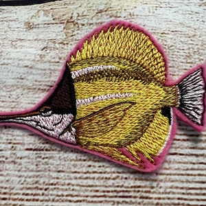May include: A yellow and brown embroidered fish patch with pink backing. The fish has a long snout and a tail fin.