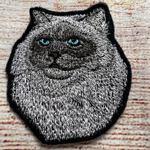 May include: Embroidered iron-on patch of a fluffy gray cat with blue eyes. The cat is looking directly at the viewer.