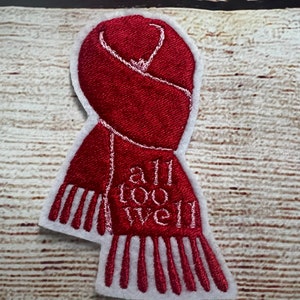 Puede incluir: Parche rojo bordado con forma de bufanda con costuras blancas. El texto "all too well" está bordado en la bufanda.