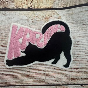 May include: Embroidered patch showing a black cat stretching, with the word "KARMA" in pink text. The patch is on a white background, set against a light brown wooden surface.