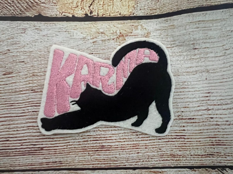 May include: Embroidered patch showing a black cat stretching, with the word "KARMA" in pink text. The patch is on a white background, set against a light brown wooden surface.