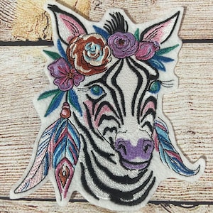 Könnte beinhalten: Gestickter Bügel-Patch mit einem Zebra mit einer Blumenkrone und Federn. Das Zebra ist weiß mit schwarzen Streifen und hat eine lila Nase. Die Blumen sind rosa, lila und orange.