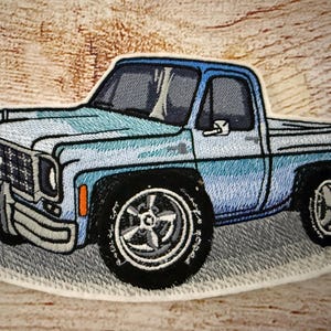 May include: Embroidered patch depicting a light blue pickup truck with black tyres and silver rims. The truck has a detailed design with a light blue body and a dark blue cab. The patch is outlined in white.