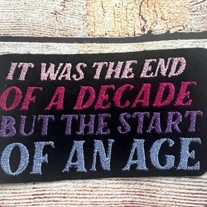 Puede incluir: Parche bordado negro con el texto "It was the end of a decade but the start of an age" en letras rosa, morado y azul.
