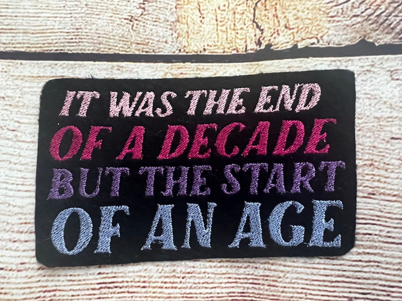 Puede incluir: Parche bordado negro con el texto "It was the end of a decade but the start of an age" en letras rosa, morado y azul.
