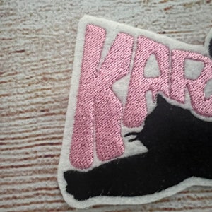 May include: Embroidered patch featuring the name "KARMA" in pink thread on a white felt backing. A black cat silhouette is integrated into the design. The patch has a neat, finished edge, suitable for application on fabric.