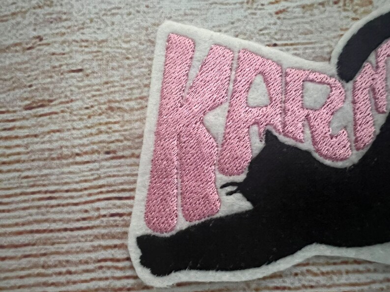 May include: Embroidered patch featuring the name "KARMA" in pink thread on a white felt backing. A black cat silhouette is integrated into the design. The patch has a neat, finished edge, suitable for application on fabric.