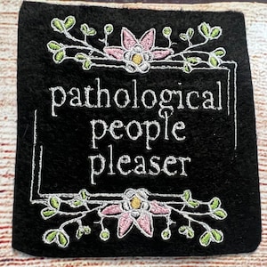 May include: Black felt patch with white embroidery that reads "pathological people pleaser". The text is framed by a white border and two pink flowers with green leaves.