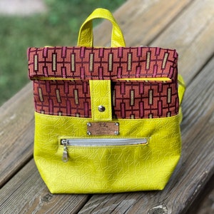 May include: A yellow faux leather backpack with a geometric pattern. The top flap is made of a red and gold patterned fabric. The backpack has a zippered pocket on the front.
