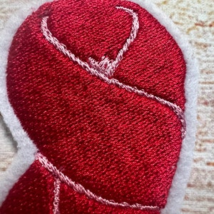 Puede incluir: Parche de fieltro rojo en forma de coraz&oacute;n con costuras blancas alrededor de los bordes. El coraz&oacute;n est&aacute; doblado y cosido en su lugar, creando un efecto 3D.