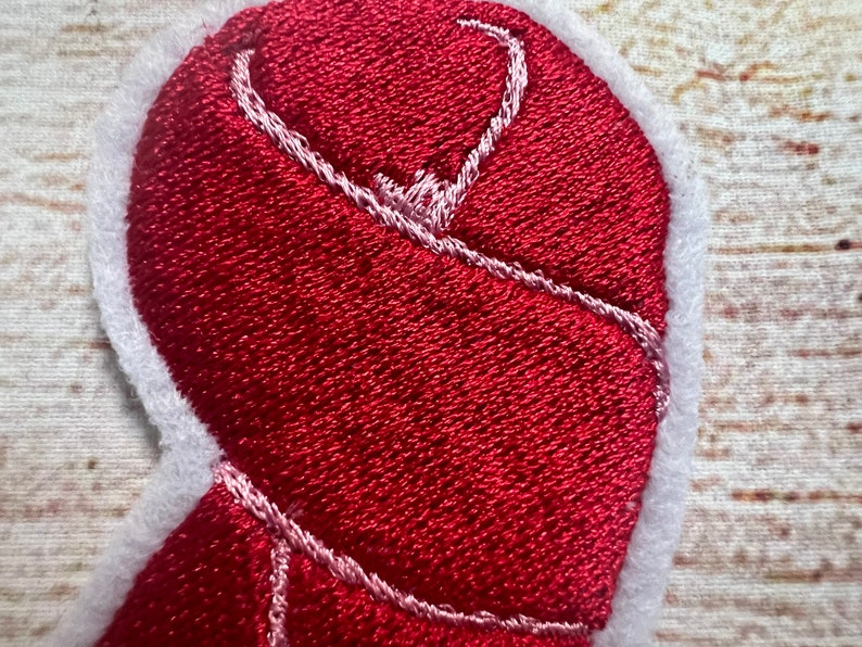 Puede incluir: Parche de fieltro rojo en forma de coraz&oacute;n con costuras blancas alrededor de los bordes. El coraz&oacute;n est&aacute; doblado y cosido en su lugar, creando un efecto 3D.