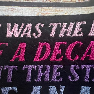 Puede incluir: Parche bordado con el texto "It was the end of a decade but the start of something new". El texto est&aacute; en hilo rosa, morado y azul sobre un fondo negro.