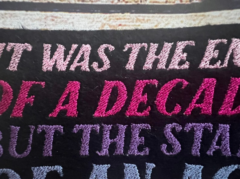 Puede incluir: Parche bordado con el texto "It was the end of a decade but the start of something new". El texto est&aacute; en hilo rosa, morado y azul sobre un fondo negro.