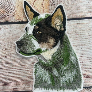 May include: Embroidered patch featuring a Blue Heeler dog in profile. The patch showcases the dog's head and upper body, with detailed stitching in shades of green, brown, white, and black. The dog's ear tips are a golden color.