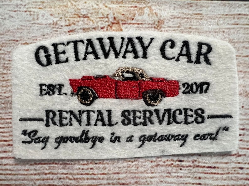 May include: Embroidered patch with a white background and black text that reads "GETAWAY CAR EST 2017 -RENTAL SERVICES- "Say goodbye in a getaway car!"  A red vintage car with a white top is embroidered in the centre of the patch.