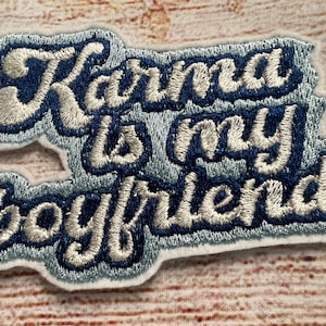 Könnte beinhalten: Ein blauer und weißer gestickter Aufnäher mit dem Text "Karma is my boyfriend" in einer kursivschrift.