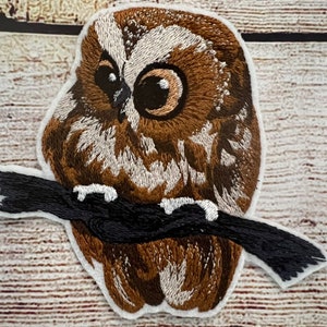 Peut inclure: Écusson brodé représentant un hibou brun et blanc perché sur une branche noire. Le hibou a de grands yeux sombres et se détache sur un fond blanc.