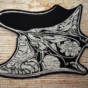 以下が含まれることがあります： 白のフローラルとリーフのデザインが施された黒色の刺繍パッチ。パッチは、湾曲した上部と尖ったエッジを持つユニークな形状をしています。複雑な刺繍が、詳細で質感のある外観を作り出しています。