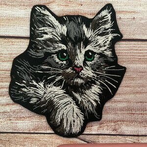 May include: Embroidered patch of a black and white cat with green eyes. The cat is looking directly at the viewer with its head tilted slightly to the side.