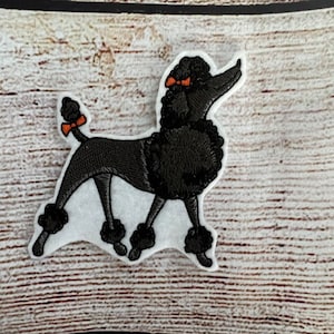 May include: Black embroidered poodle patch with orange bows. The poodle is standing on its hind legs with its front paws raised. The patch is on a white background.