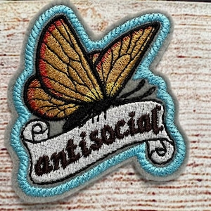 May include: An embroidered iron-on patch with a blue border. A yellow and orange butterfly with brown accents is flying above a white scroll with the text "antisocial" in brown lettering.
