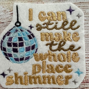 May include: A white felt patch with gold embroidery that reads "I can still make the whole place shimmer". A blue and purple disco ball hangs from the top of the patch.
