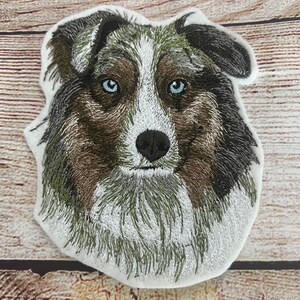 Op de afbeelding: Gestikte patch van de kop van een Australian Shepherd hond in blue merle. De hond heeft bruine vacht, blauwe ogen en een witte vlek op zijn borst.