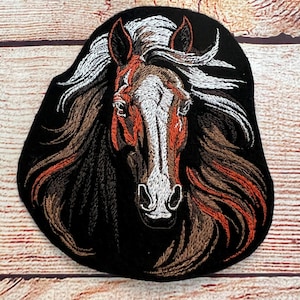 Op de afbeelding: Geborduurde patch met een paardenkop met een vloeiende manen. Het paard heeft een wit gezicht met bruine en oranje details, tegen een zwarte achtergrond. De manen zijn een mix van witte, bruine en zwarte draden, wat een gedetailleerd ontwerp creëert.