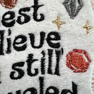Puede incluir: Parche bordado con el texto "I best believe I'm still loved". El parche es blanco con bordado negro y detalles de purpurina dorada y plateada.