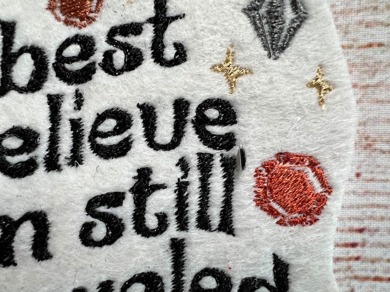 Puede incluir: Parche bordado con el texto "I best believe I'm still loved". El parche es blanco con bordado negro y detalles de purpurina dorada y plateada.