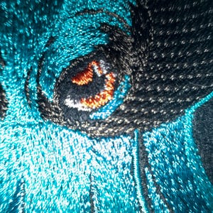 Puede incluir: Primer plano de un diseño bordado con un ojo detallado. El ojo tiene una pupila negra con acentos naranjas y marrones, rodeada de pespuntes negros y plateados. El fondo es de un color turquesa vibrante.