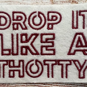 May include: A white fabric patch with the text "DROP IT LIKE A THOTTY" embroidered in maroon thread.