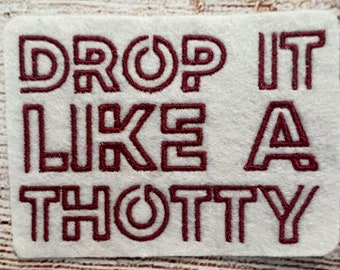 Puede incluir: Un parche de tela blanco con el texto "DROP IT LIKE A THOTTY" bordado en hilo marrón.