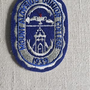 May include: A blue and silver embroidered patch with the text "MOUNT ALOYSIUS JUNIOR COLLEGE" and the year "1939". The patch has a scalloped edge and features a central design of a building and a lamp.