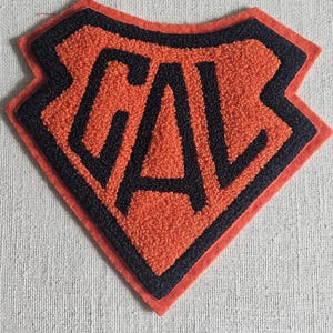 May include: Orange and black felt patch with the letters "CAL" in a bold, blocky font. The patch has a diamond shape with a black border and a textured orange center. The letters are black.