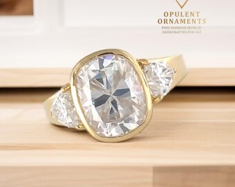 May include: A gold ring with a large, oval-cut, clear gemstone in the center, flanked by two smaller, triangular-cut gemstones. The ring is set on a light-colored wooden surface. The text "OPULENT ORNAMENTS" is visible.