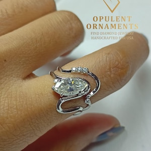 May include: A silver ring with a marquise-cut diamond and smaller accent stones. The ring wraps around the finger in a unique, modern design. The text "OPULENT ORNAMENTS" and "FIND DIAMOND JEWELRY HANDCRAFTED FOR USA" is visible.