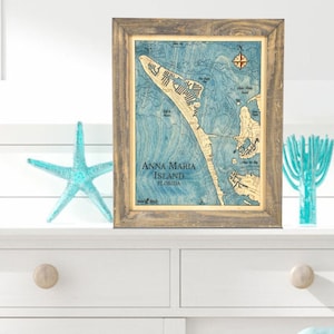 May include: A framed wooden map of Anna Maria Island, Florida, in shades of blue and beige. The map is displayed on a white dresser, alongside a blue starfish and a decorative glass piece. The frame has a rustic, weathered appearance.