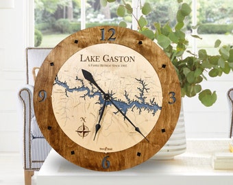 May include: A wooden clock with a map of Lake Gaston, North Carolina. The clock face is brown with black numbers and hands. The map is beige with blue water features. The text "Lake Gaston A Family Retreat Since 1958" is printed on the clock face.