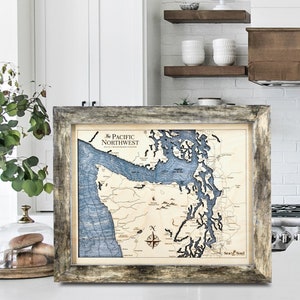 May include: A framed wooden map of the Pacific Northwest with a blue and brown color scheme. The map includes the text "The Pacific Northwest" and a compass rose. The frame is a weathered gray color.