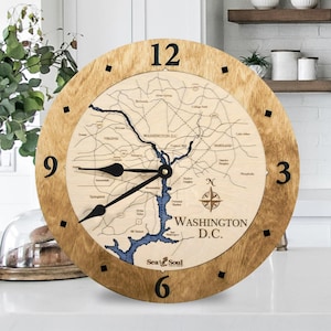 May include: A round wooden clock featuring a map of Washington D.C. The clock face displays the city's layout with blue water features and black hands. The clock has a light brown wooden frame with black markers at the hour positions.