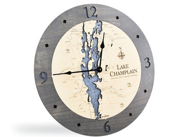 May include: A gray wooden wall clock with a map of Lake Champlain in the center. The clock face is surrounded by a gray wooden frame with black numbers and hands. The map is in shades of blue and brown, with the lake highlighted in blue. The clock has a compass rose in the lower right corner.